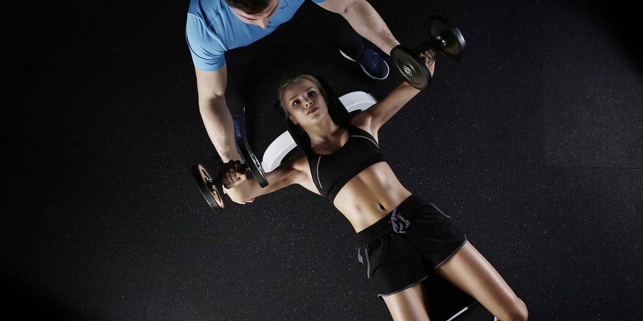 Female athlete performing dumbbell chest press with trainer assistance.